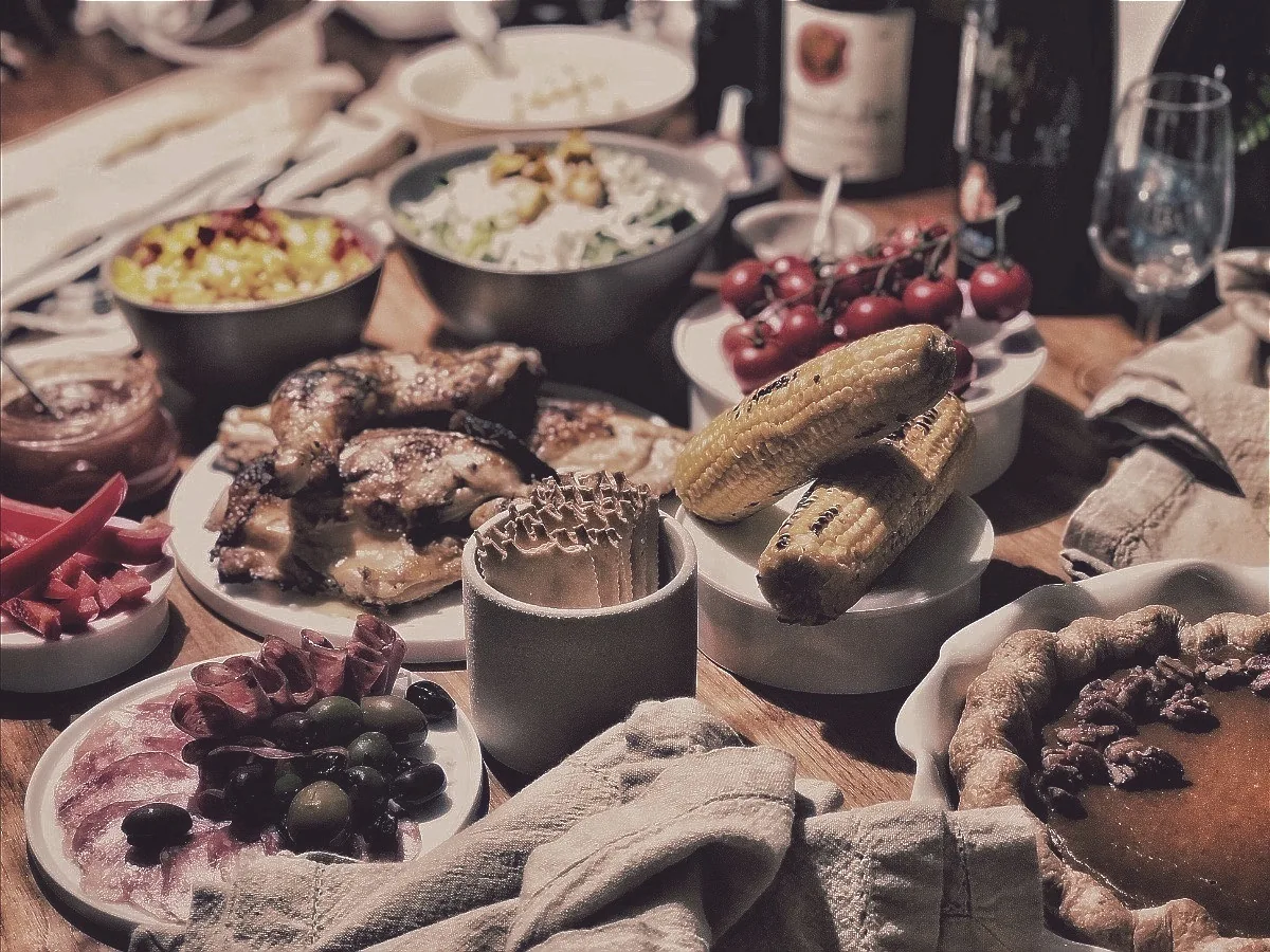 A crowded Thanksgiving table with roast bird, grilled corn, charcuterie, olives, pecan pie and open wine